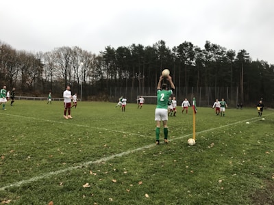 A group of soccer players is engaged in a match on a grassy field, surrounded by tall trees. One player wearing a green jersey is preparing to throw the ball in from the sideline. Other players in white and green jerseys are positioned across the field, ready to resume play.