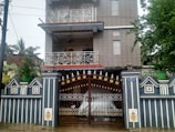 A two-story house with decorative metal railings on the balconies and a large, ornate metal gate at the entrance. The facade is covered with patterned tiles, and potted plants are placed on the boundary wall and balconies. A sign is visible near the entrance. The setting includes some greenery with trees on the side.
