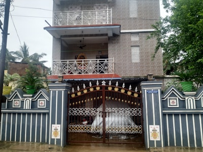 A two-story house with decorative metal railings on the balconies and a large, ornate metal gate at the entrance. The facade is covered with patterned tiles, and potted plants are placed on the boundary wall and balconies. A sign is visible near the entrance. The setting includes some greenery with trees on the side.