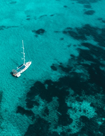 A top-down view of a sailboat floating on crystal-clear turquoise water with dark patches that suggest reefs or seaweed. The boat is anchored or calmly drifting, offering a serene coastal setting.