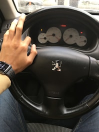 A person’s hand is resting on a car steering wheel with a logo at the center. The speedometer and other gauges are visible in the dashboard. The person is wearing a smartwatch and blue jeans. The steering wheel appears to be leather, and the airbag label is visible.