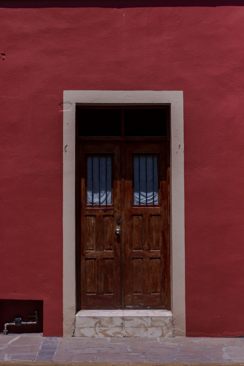 The image shows a red-painted building with two large wooden doors, one on each side, situated at an angle and slightly open. The doors are framed by a window above them, which is currently closed. A small portion of the ground can be seen in front of the door, suggesting that it might be located near a sidewalk or entrance to a building.

The red paint on the walls creates a striking contrast with the wooden doors and windows, making the scene visually appealing. The positioning of the doors at an angle adds depth to the image, giving it a three-dimensional feel.