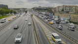 A busy multi-lane highway with heavy traffic, including a mix of cars, taxis, and buses. Surrounding the road are high-rise buildings and advertisements on a retaining wall. The sky is overcast, adding a grayish tone to the scene.