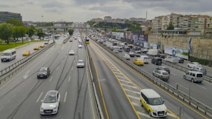 A busy multi-lane highway with heavy traffic, including a mix of cars, taxis, and buses. Surrounding the road are high-rise buildings and advertisements on a retaining wall. The sky is overcast, adding a grayish tone to the scene.
