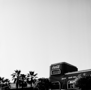 A black and white image features a building with Coca-Cola signage prominently displayed. Palm trees are visible in the foreground, enhancing the contrast against the clear sky.