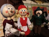 A group of intricately designed marionette puppets with cheerful facial expressions. The puppets feature vibrant costumes, with one dressed in a red outfit, another in a green suit with a hat, and a third with grey hair and a traditional vest. They are set in a theatrical backdrop, suggesting a performance or storytelling scenario.