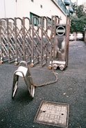 A metallic expandable gate is positioned across an asphalt surface, with part of it chained to a nearby metal barrier. The background shows the side of a building with windows and parked cars. The ground also displays a utility cover with engraved markings.