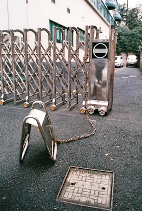 A metallic expandable gate is positioned across an asphalt surface, with part of it chained to a nearby metal barrier. The background shows the side of a building with windows and parked cars. The ground also displays a utility cover with engraved markings.