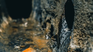 A close-up of a rugged, textured surface with a narrow channel running through it. Water is seen flowing gently along the channel, creating a glossy reflection on the rough, earthy surface. The scene captures a sense of nature, with the focus on the interaction between the solid, rocky textures and the flowing water.