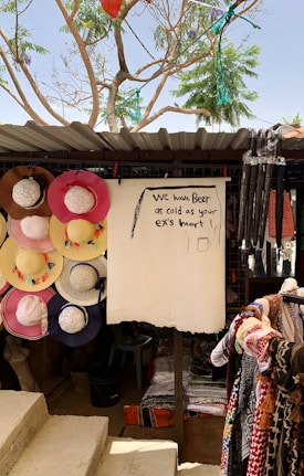 A market stall displaying a variety of colorful sun hats hanging on the left side and patterned textiles on the right. A humorous sign in the center reads 'We have beer as cold as your ex's heart,' accompanied by simple drawings. Above, a corrugated roof and a tree with green foliage provide shade.