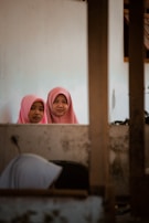 Two young women wearing pink hijabs are standing behind a wall and looking directly at the camera. The setting appears to be indoors, with another person, partially visible in the foreground, seemingly engaged in a different activity. The indoor environment appears modest and somewhat aged, with visible wear on the walls.
