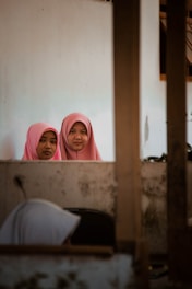 Two young women wearing pink hijabs are standing behind a wall and looking directly at the camera. The setting appears to be indoors, with another person, partially visible in the foreground, seemingly engaged in a different activity. The indoor environment appears modest and somewhat aged, with visible wear on the walls.