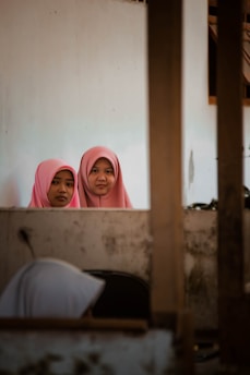 Two young women wearing pink hijabs are standing behind a wall and looking directly at the camera. The setting appears to be indoors, with another person, partially visible in the foreground, seemingly engaged in a different activity. The indoor environment appears modest and somewhat aged, with visible wear on the walls.