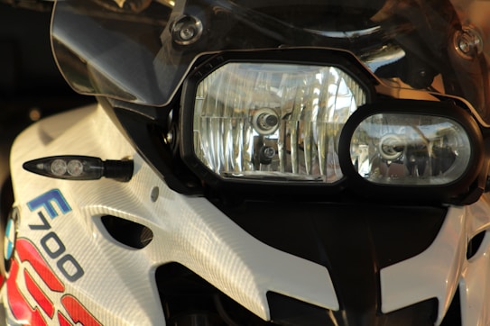 Close-up view of a motorcycle's front, focusing on the headlight assembly, which includes two distinct lights. The motorcycle is adorned with stickers and labels, including an 'F700' insignia. The color scheme is predominantly white with black accents.