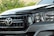 Close-up view of a car's front grille and headlight, featuring a prominent logo. The car's exterior is clean and well-maintained, with reflections visible on the glossy surface. Greenery is visible in the background.
