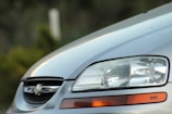 Close-up of the front portion of a silver car, focusing on the headlight and part of the grille. The car features a Chevrolet emblem on the grille, and the headlight is detailed with reflections and clear lenses. The background is blurred with hints of greenery.