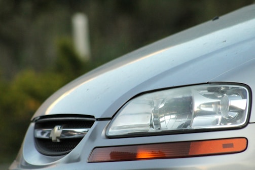 Close-up of the front portion of a silver car, focusing on the headlight and part of the grille. The car features a Chevrolet emblem on the grille, and the headlight is detailed with reflections and clear lenses. The background is blurred with hints of greenery.