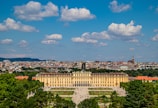 An impressive historical palace stands prominently in the center with a sprawling garden in front. The structure is a long, yellow building with numerous windows and a symmetrical design. Visitors are scattered across the open area, forming a bustling scene. In the background, a cityscape stretches across the horizon with a clear blue sky dotted with fluffy clouds overhead.