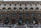 A historic building showcases elaborate architectural details such as ornate stone carvings and multiple arched windows across several floors. The facade combines both lighter and darker textured stones, with a series of large, symmetrical windows framed by decorative columns.