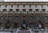 A historic building showcases elaborate architectural details such as ornate stone carvings and multiple arched windows across several floors. The facade combines both lighter and darker textured stones, with a series of large, symmetrical windows framed by decorative columns.