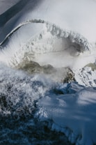 A frozen landscape with snow-covered surfaces and flowing water partially shrouded in ice. The textures are rough and jagged, with visible layers of compacted snow, creating a dynamic and cold environment.
