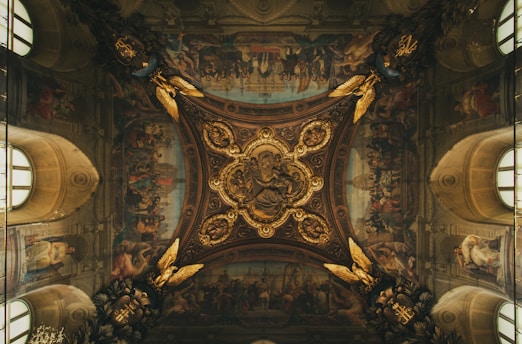 An intricately decorated ceiling features elaborate frescoes with religious and historical imagery, surrounded by ornate golden embellishments and detailed artwork. The central medallion is richly adorned with carved figures and gold leaf, displaying a masterful blend of art and architecture.