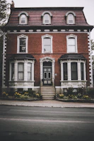 A stately brick house with three floors and a distinct architectural style featuring arched windows. The facade includes decorative trim, and a central entrance is accessed by a set of stairs. Neatly maintained gardens with yellow flowers border the pathway leading to the door.
