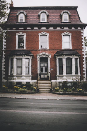 A stately brick house with three floors and a distinct architectural style featuring arched windows. The facade includes decorative trim, and a central entrance is accessed by a set of stairs. Neatly maintained gardens with yellow flowers border the pathway leading to the door.