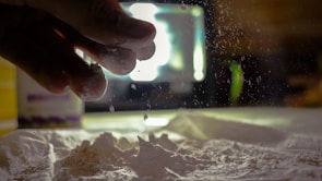 A close-up of fingers sprinkling fine white powder onto a surface, captured in motion with some particles suspended in the air. The background features a blurry light source and container, creating a dramatic and moody atmosphere.