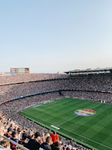 An expansive soccer stadium filled with thousands of spectators. The green field in the center features players and markings, surrounded by vibrant stands and advertising boards on the upper sides of the stadium. The audience is actively watching the game.