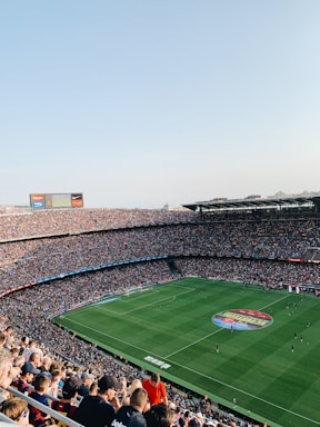 An expansive soccer stadium filled with thousands of spectators. The green field in the center features players and markings, surrounded by vibrant stands and advertising boards on the upper sides of the stadium. The audience is actively watching the game.
