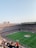An expansive soccer stadium filled with thousands of spectators. The green field in the center features players and markings, surrounded by vibrant stands and advertising boards on the upper sides of the stadium. The audience is actively watching the game.