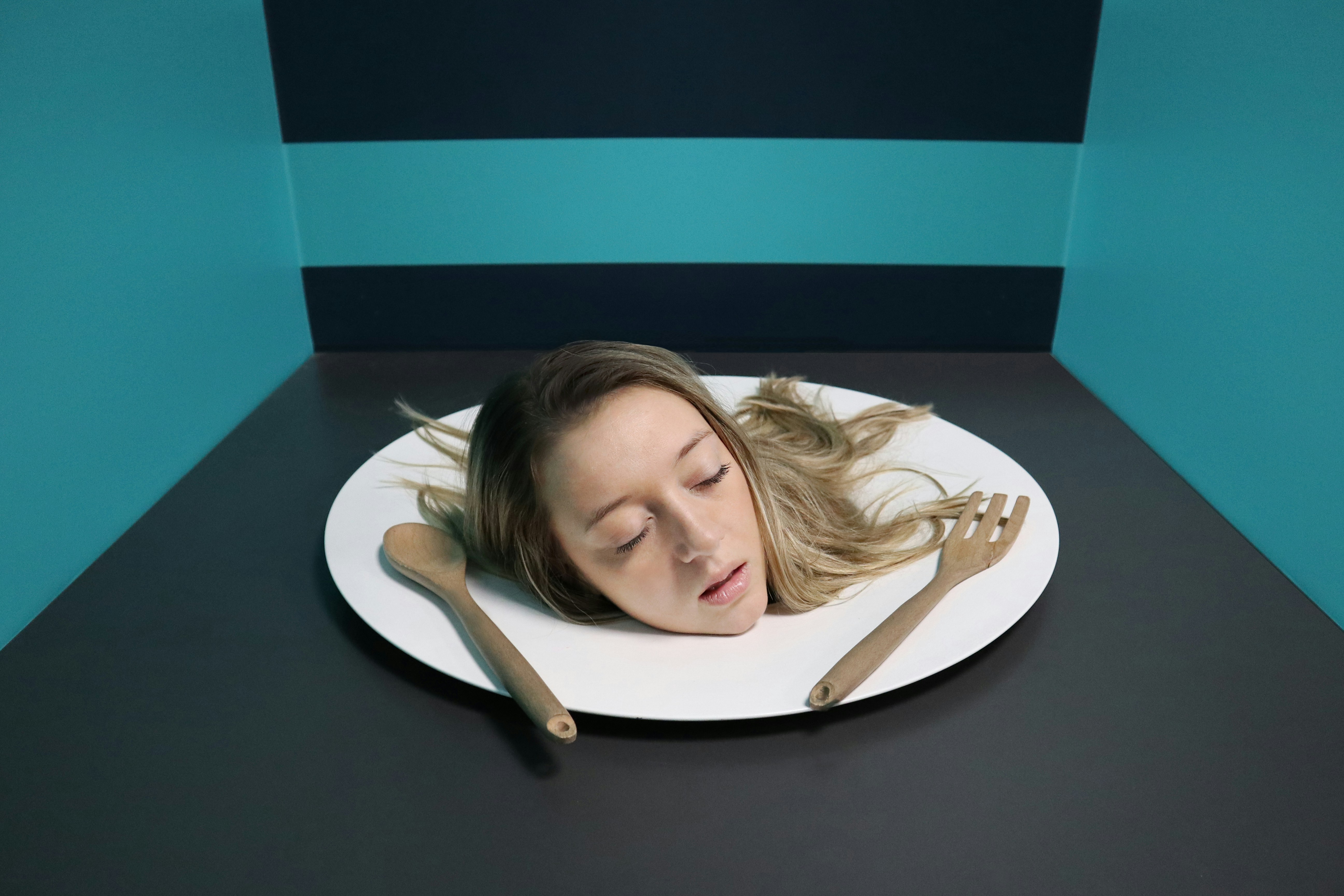 Person with long hair seated in a small blue room with their head poking through a circular cutout in a table, hands resting on the table, symbolizing entrapment and denial.