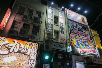 A vibrant urban streetscape at night with brightly illuminated commercial signs and advertisements covering the facade of a multi-story building. The signs feature colorful graphics and Japanese text, promoting various businesses and products. The scene is lively and bustling, giving a sense of a busy city area.