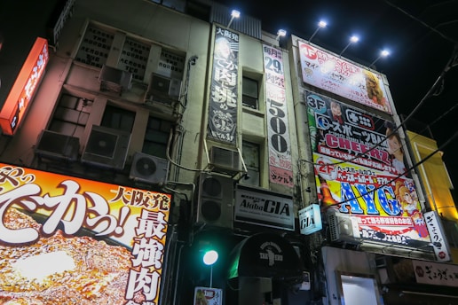 A vibrant urban streetscape at night with brightly illuminated commercial signs and advertisements covering the facade of a multi-story building. The signs feature colorful graphics and Japanese text, promoting various businesses and products. The scene is lively and bustling, giving a sense of a busy city area.