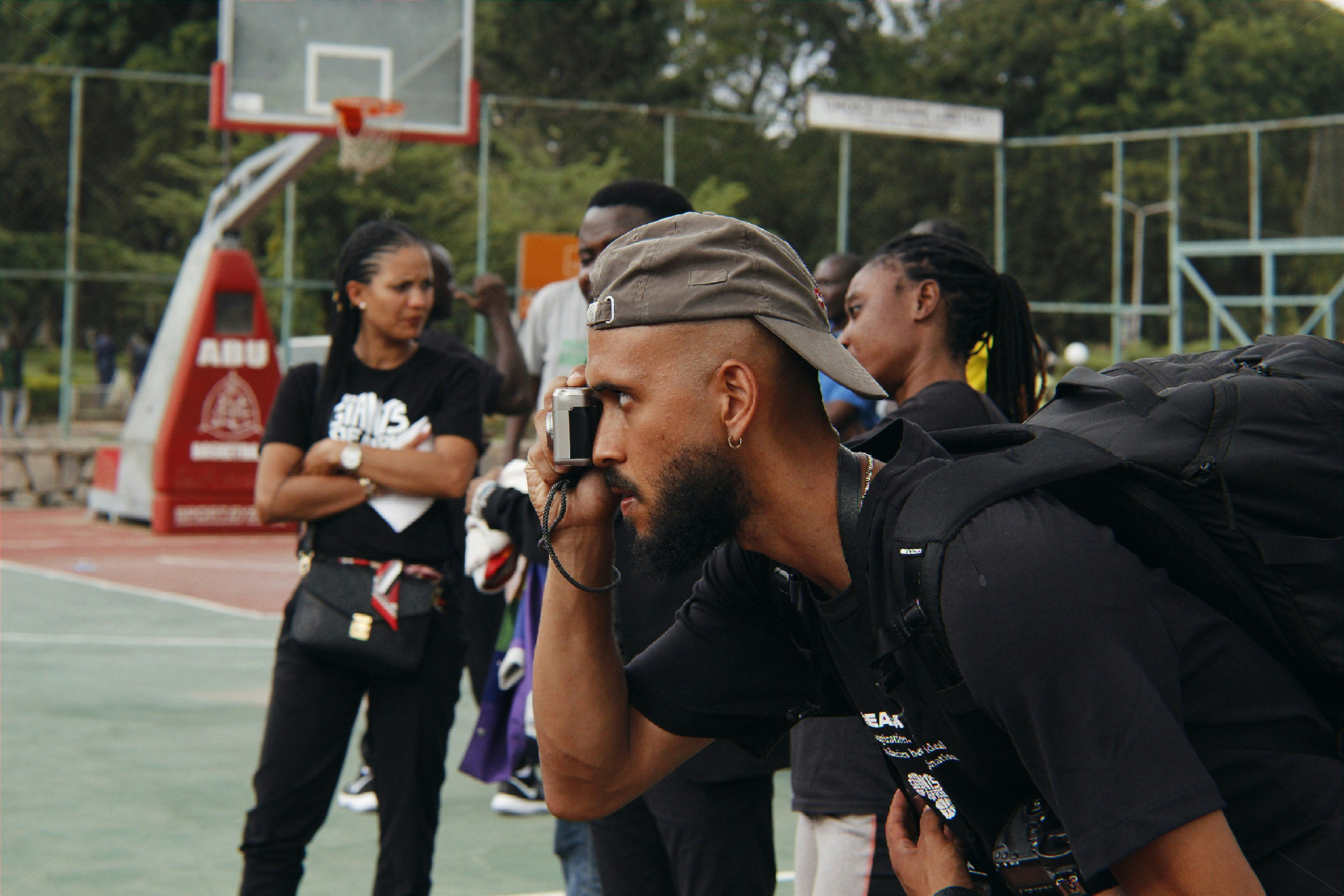 A group of people gather on an outdoor basketball court. A man in the foreground holds a camera up to his eye, wearing a cap backward and a backpack. Other individuals stand in the background, some appearing to chat and observe the scene. A basketball hoop and a scoreboard are visible, surrounded by a metal fence and trees.