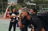 A group of people gather on an outdoor basketball court. A man in the foreground holds a camera up to his eye, wearing a cap backward and a backpack. Other individuals stand in the background, some appearing to chat and observe the scene. A basketball hoop and a scoreboard are visible, surrounded by a metal fence and trees.
