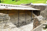 An ancient archaeological site features partially excavated stone walls and structures. The area is covered with a protective corrugated metal roof supported by metal beams. The stonework displays a variety of textures and patterns, with some areas showing erosion or damage. Surrounding the site, there are lush green areas indicating a rural or remote location.