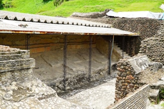 An ancient archaeological site features partially excavated stone walls and structures. The area is covered with a protective corrugated metal roof supported by metal beams. The stonework displays a variety of textures and patterns, with some areas showing erosion or damage. Surrounding the site, there are lush green areas indicating a rural or remote location.