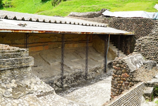 An ancient archaeological site features partially excavated stone walls and structures. The area is covered with a protective corrugated metal roof supported by metal beams. The stonework displays a variety of textures and patterns, with some areas showing erosion or damage. Surrounding the site, there are lush green areas indicating a rural or remote location.