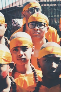 A group of young individuals wearing bright orange clothing and headbands, with shaved heads and decorative beads. Some have glasses, adding a modern touch to their traditional appearance. They appear in a warm, sunlit setting.
