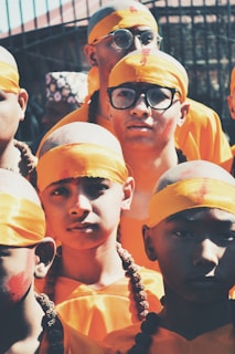 A group of young individuals wearing bright orange clothing and headbands, with shaved heads and decorative beads. Some have glasses, adding a modern touch to their traditional appearance. They appear in a warm, sunlit setting.