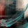 A dynamic brushstroke of teal paint sweeps diagonally across a reflective surface, with blurred reflections of a building in the background. The combination of colors and abstract form creates a sense of movement and fluidity.