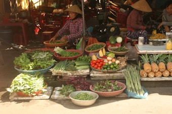 A bustling market scene with a variety of fresh vegetables and fruits displayed. A vendor wearing a conical hat is preparing produce for sale. The assortment includes leafy greens, cucumbers, tomatoes, limes, pineapples, and herbs, all arranged in baskets and trays. The setting appears to be outdoors under partial shade from a canopy.