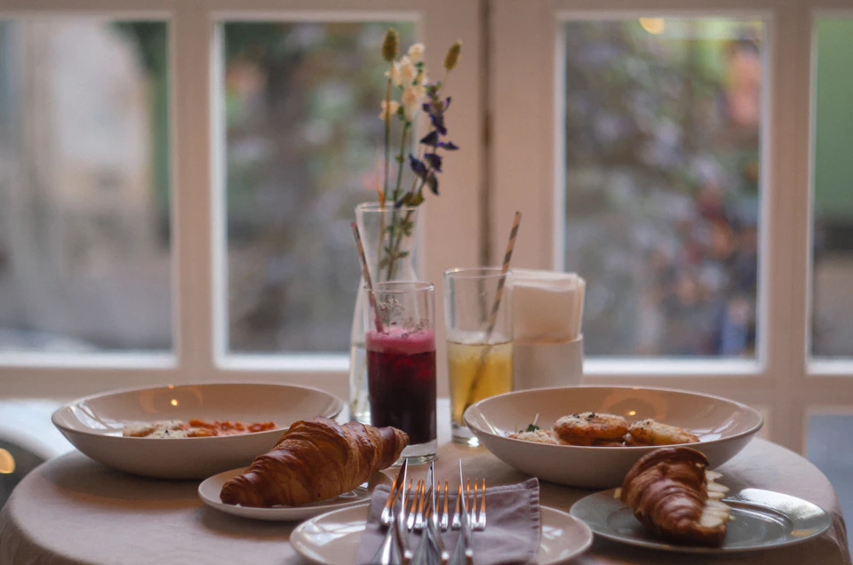The image depicts a table set for two with plates of food and drinks, including croissants, juice, and water. The table is positioned near a window that offers a view of trees outside. A vase filled with flowers adds a touch of nature to the scene. Two forks are placed on one side of the table, ready to be used by the diners.