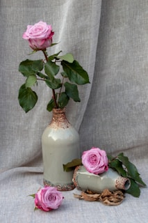 Pink roses are arranged aesthetically with one standing upright in a tall ceramic vase, while two more roses rest on the side, surrounded by some leaves and dried petals. The backdrop is a linen fabric that complements the muted tones of the vases and the vibrant shade of the flowers.