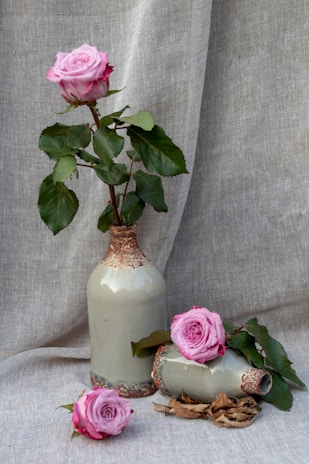 Pink roses are arranged aesthetically with one standing upright in a tall ceramic vase, while two more roses rest on the side, surrounded by some leaves and dried petals. The backdrop is a linen fabric that complements the muted tones of the vases and the vibrant shade of the flowers.
