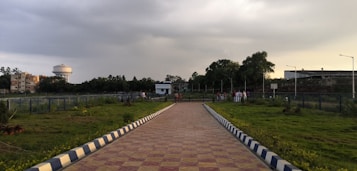 A paved walkway leading to a small white building. On either side, there's green grass with scattered plants. Several people are gathered near the building. In the background, a water tower is visible to the left, alongside some buildings, while tall trees border the area.