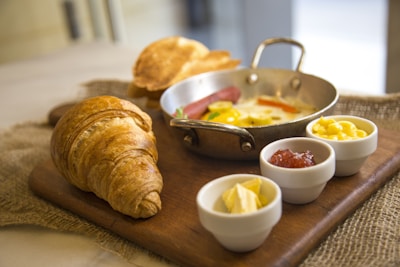 A breakfast presentation featuring a croissant, small frying pan with fried eggs and vegetables, small bowls containing butter, jam, and what appears to be cheese, all arranged on a wooden cutting board.