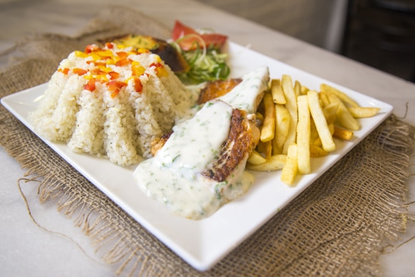 A square white plate features a serving of white rice topped with small diced red and yellow bell peppers. Next to the rice, there is a piece of grilled fish covered in a creamy white sauce. A portion of golden, crispy French fries accompanies the fish, and a small salad with lettuce and sliced tomatoes is arranged at the back. The setting is complemented by a burlap mat beneath the plate, adding a rustic touch.